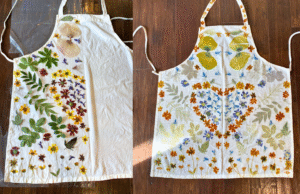 Two white aprons printed with colorful herbs and flowers. One design includes a large orange heart.