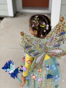 Back view of a young girl with flowers woven into her hair and dress. Wearing colorful patterned wings by artist Deena Class.