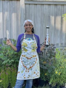Smiling woman outdoors wearing a colorful eco-printed apron and holding tools.
