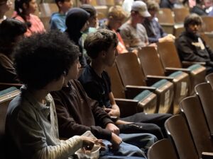 Teenagers in a school auditorium, watching the stage closely.
