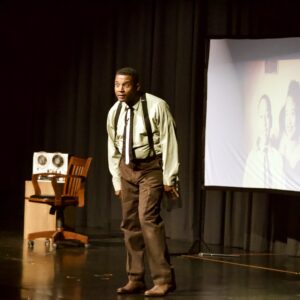 Black actor in suspenders and brown pants, onstage performing in character.