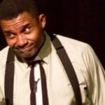 Black actor in suspenders, white shirt, and brown paints. Onstage, he gestures while speaking in character.
