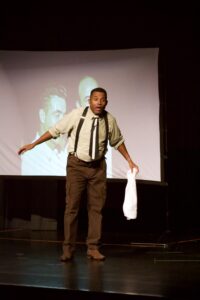 Black actor on stage in suspenders and brown pants, with a surprised face and holding a white towel.