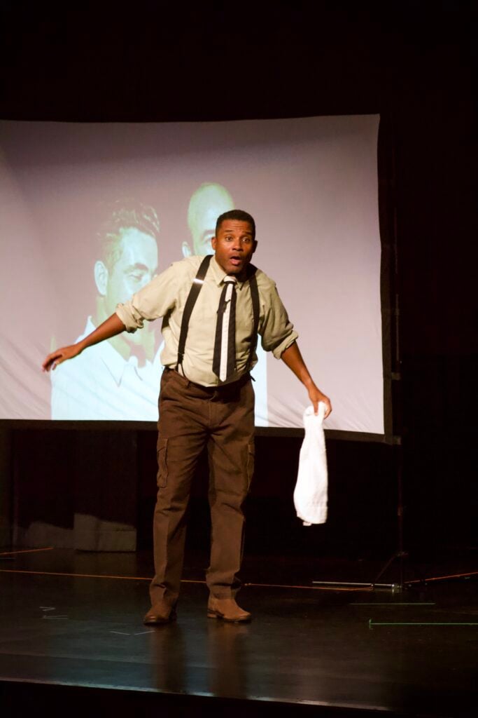 Black actor on stage in suspenders and brown pants, with a surprised face and holding a white towel.