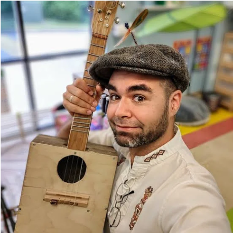 Man with beard and wearing hat holds up stringed intstrument.