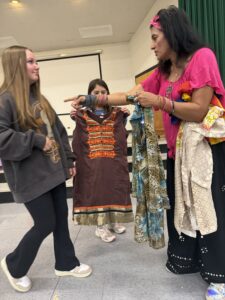 Indian woman chooses Bollywood dance costumes for seventh-grade girls as they smile.