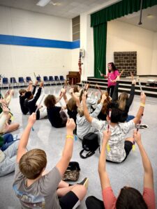 Students sit on a multipurpose room floor with hands raised to ask an Indian Bollywood dancer questions.