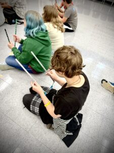 Student kneels on a school's multipurpose room floor holding colorful Indian folk dance sticks.