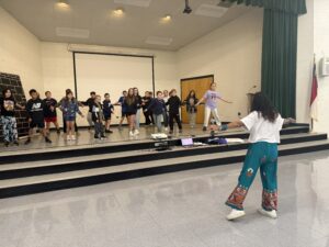Students dance on stage in a multipurpose room floor while an Indian Bollywood dancer guides them.