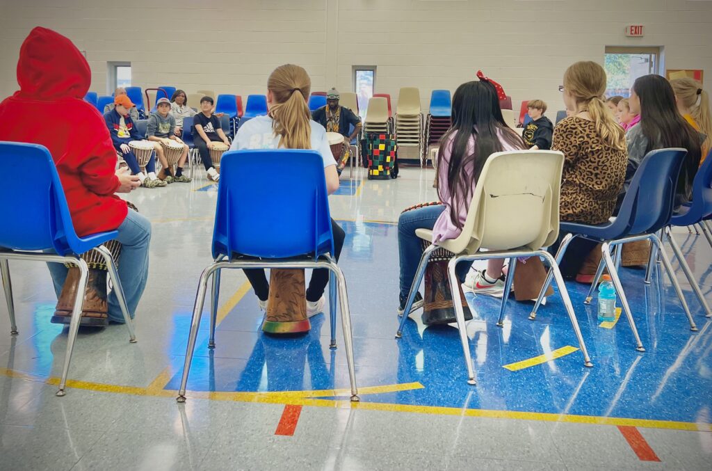 A view from behind the blue and white school chairs of students sitting in a circle, learning to play djembe drums.