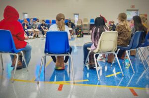 A view from behind the blue and white school chairs of students sitting in a circle, learning to play djembe drums.