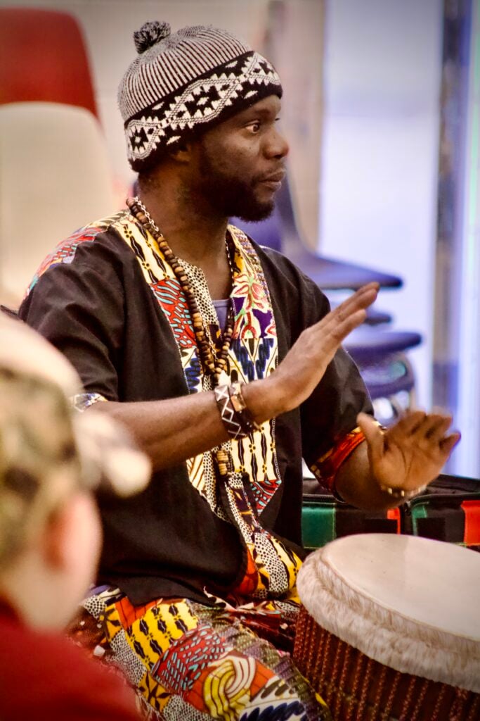 Black man in Senegalese attire playing a djembe drum.
