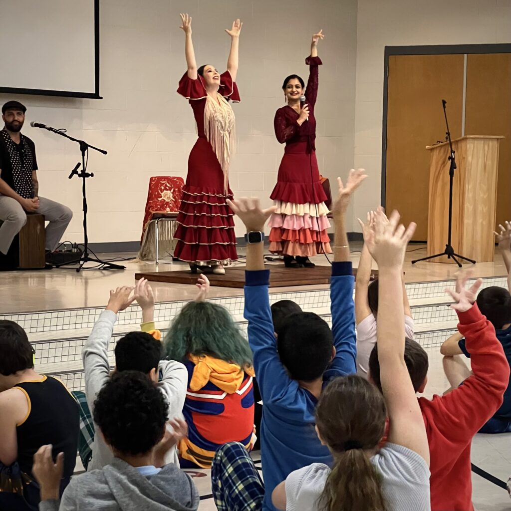 Two female Flamenco dancers perform for elementary students, who are raising their arms like the dancers.