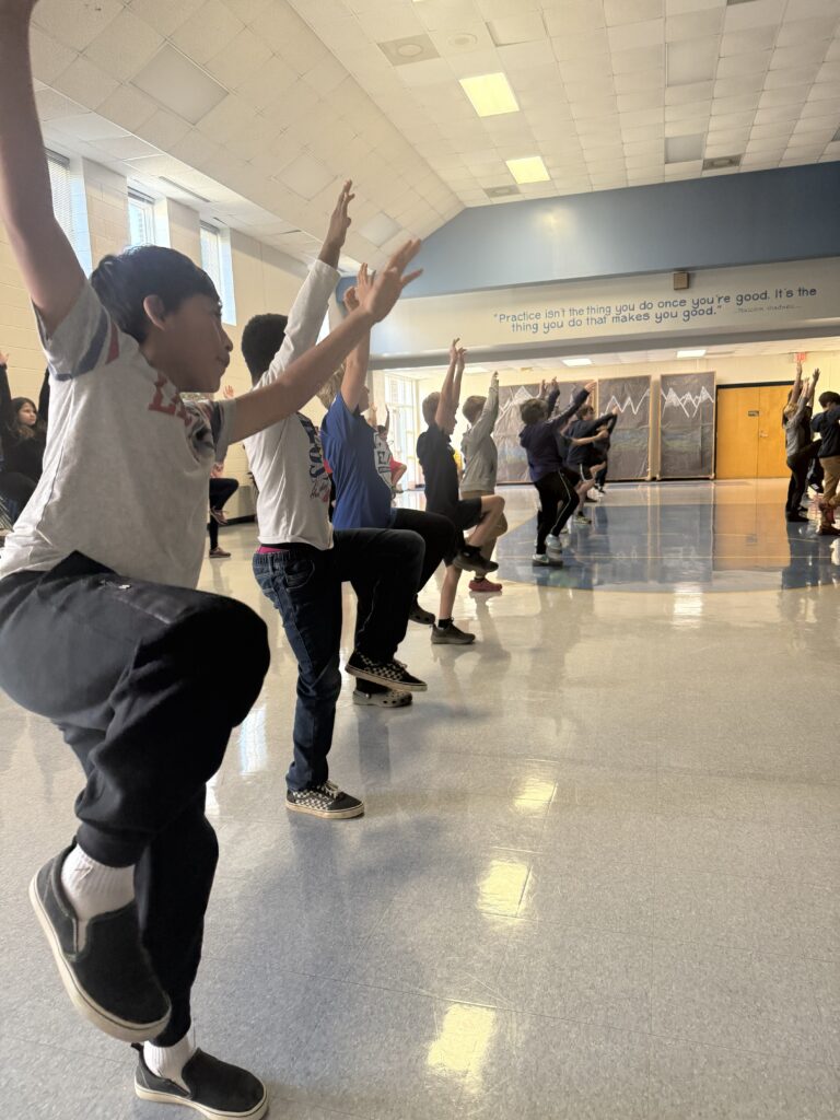 A line of middle grade students dance in a school multipurpose room.