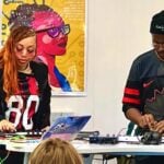 Black woman with long hair and glasses and Black man with glasses and knit cap work with digital music equipment in front of a class.