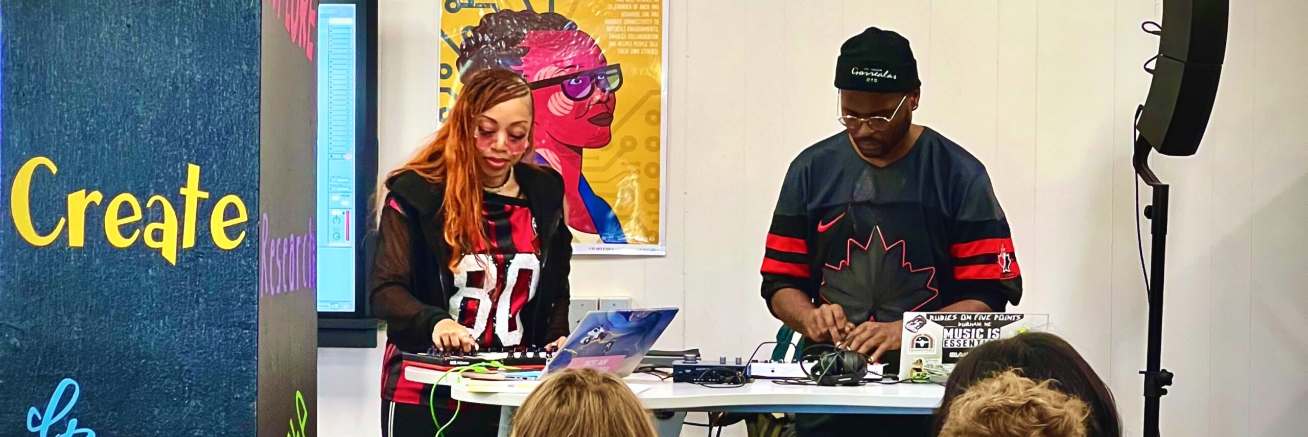 Black woman with long hair and glasses and Black man with glasses and knit cap work with digital music equipment in front of a class.