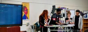 A Black woman and a Black man stand at a table helping two high schoolers use digital music equipment.