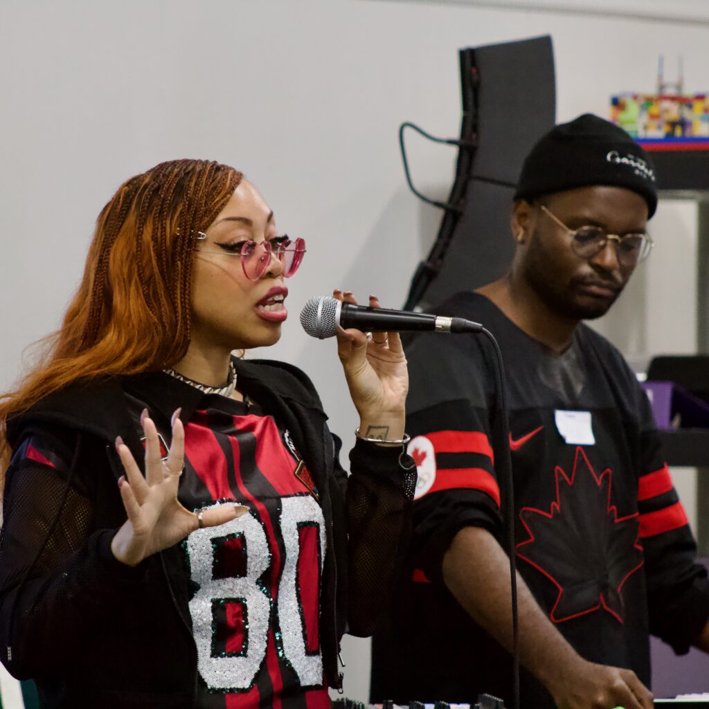 Black man in black knit cap and Black woman wearing glasses. Woman is speaking into a microphone.