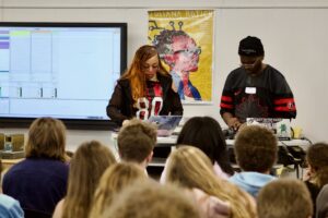 A Black woman with long hair and a Black man with a knit cap perform beats live for a school audience.