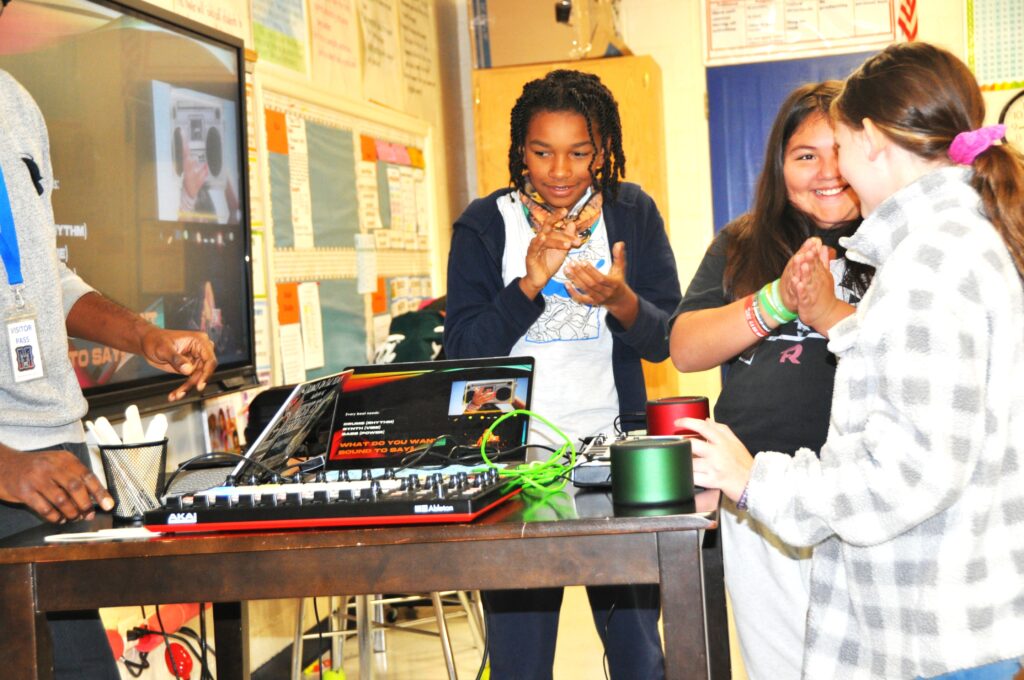A student with brown skin, a student with light brown skin, and a student with pale skin congratulate each other in front of digital beatmaking equipment.