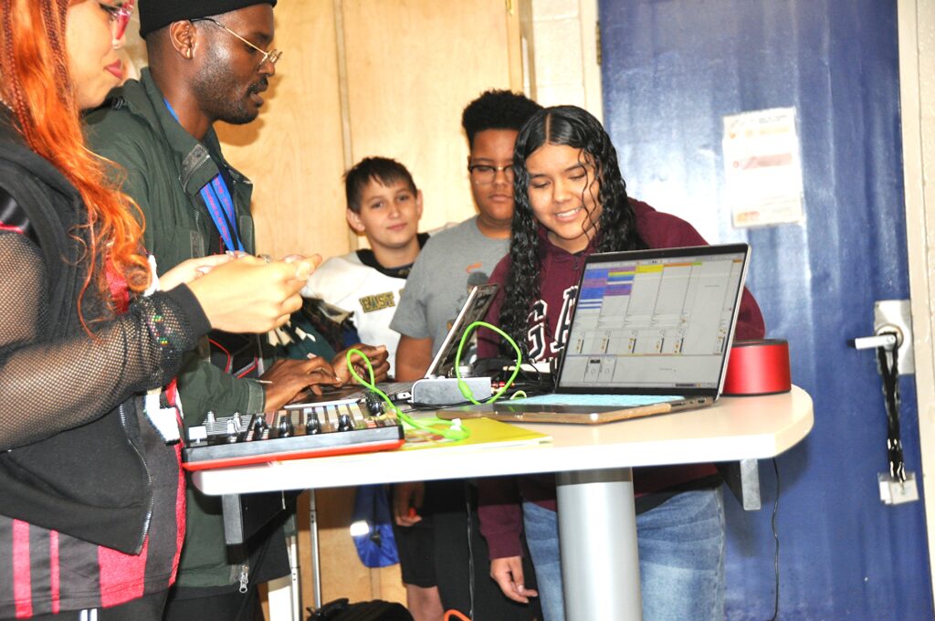 A student with light brown skin and long dark hair smiles at the front of a short line of students ready to work with digital music equipment.