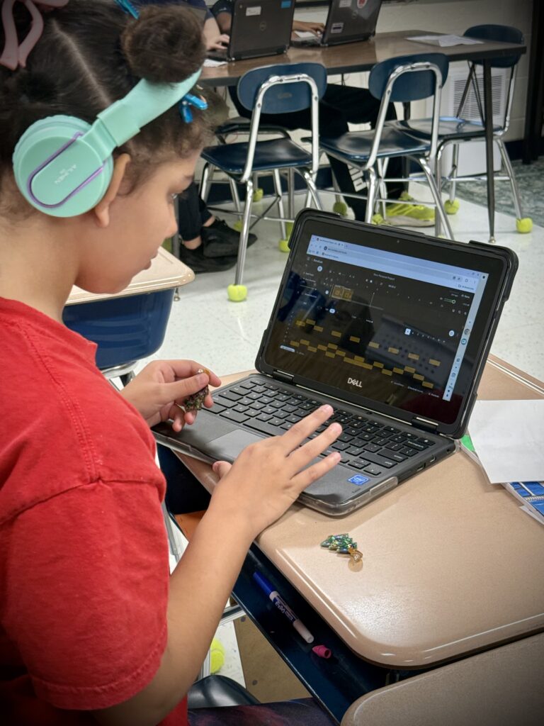 A student with light brown skin and teal headphones works at her laptop.