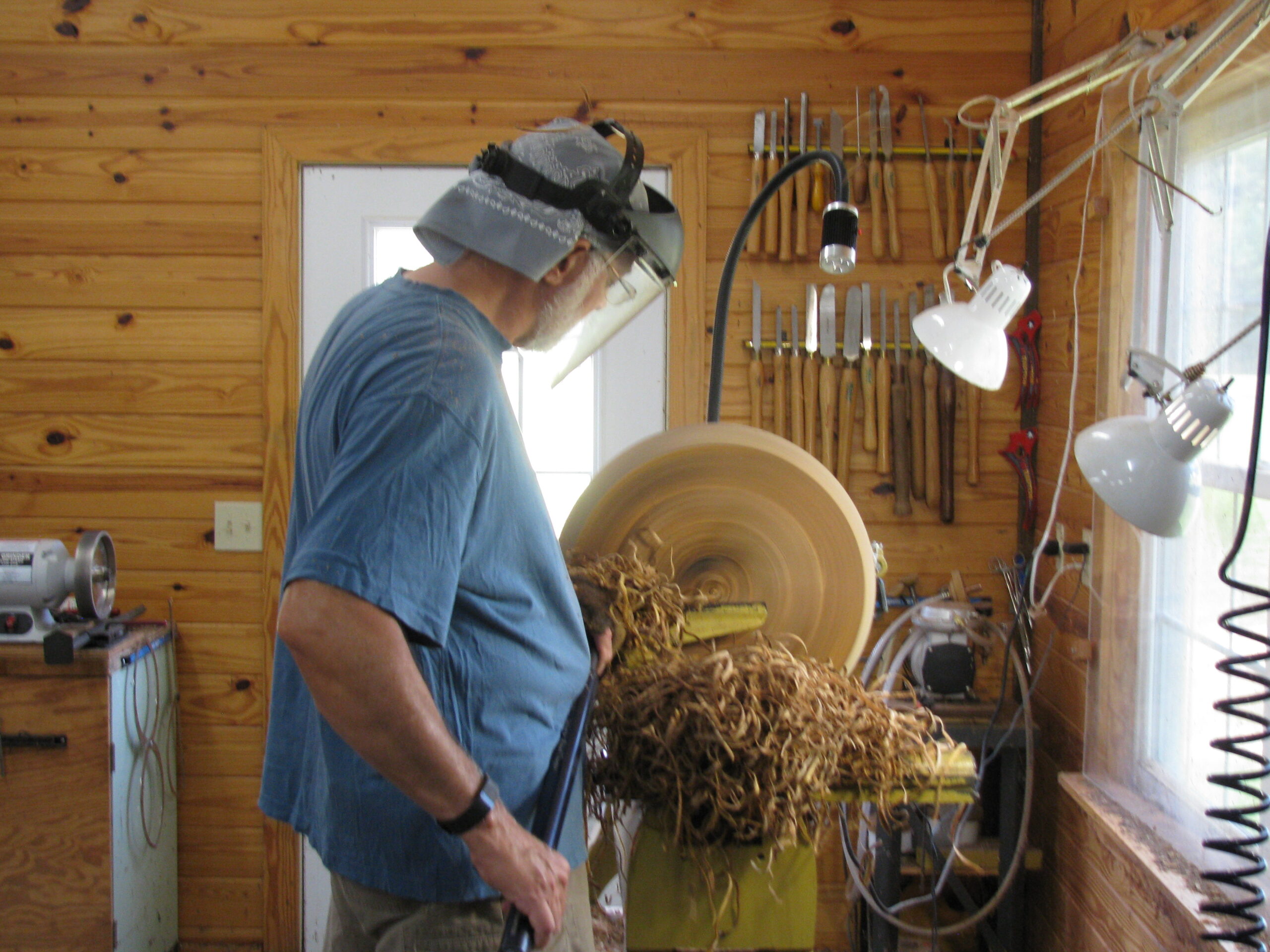 a man is woodturning and wearing a protective face shield and blue tee shirt.