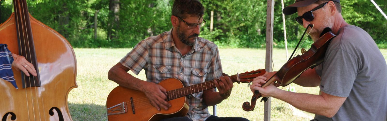 Three musicians play instruments outdoors under a tent: one plays a double bass, one plays a guitar, and one plays a violin.