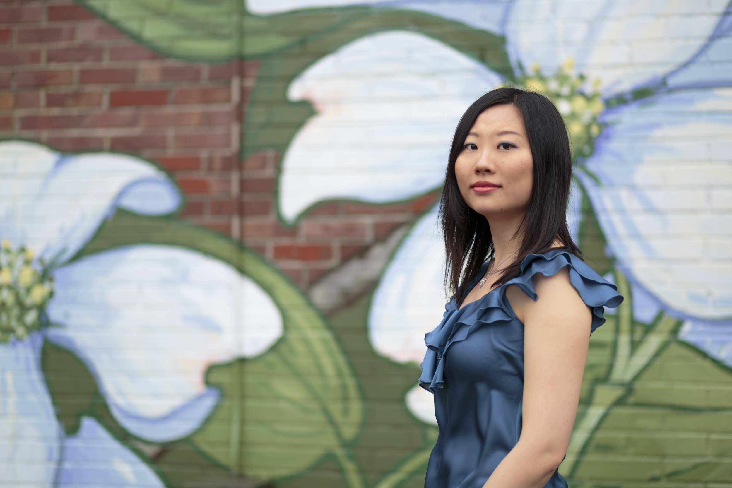 A woman of asian descent with shoulder length black hair in 3/4 profile. she is wearing a blue, sleeveless blouse and is standing in front of a mural of magnolia flowers on brick.