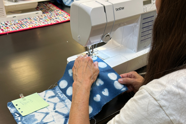 A person is sewing two pieces of blue fabric with white designs on a white Brother sewing machine.