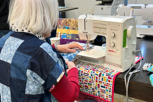 A person wearing a denim patchwork jacket sits at a sewing machine, with their hands guiding fabric under the needle.