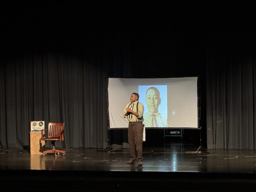 Mike Wiley on stage in white shirt and brown pants with image of Emmitt Till on the screen behind him