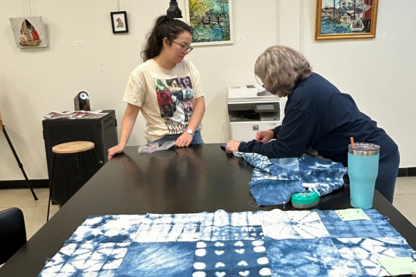 Two people are working on a long piece of blue and white tie-dye fabric laid out on a dark table. One person is standing and looking at the fabric, while the other is seated and appears to be sewing or measuring it.