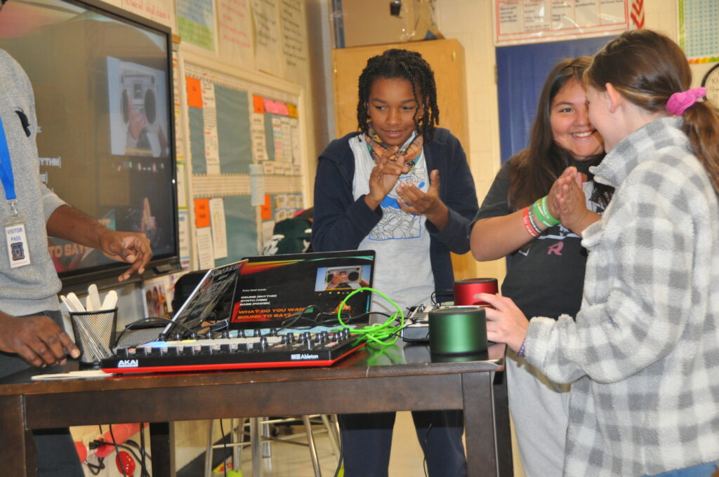 Three middle school students standing in front of beat making computers clapping their hands and smiling