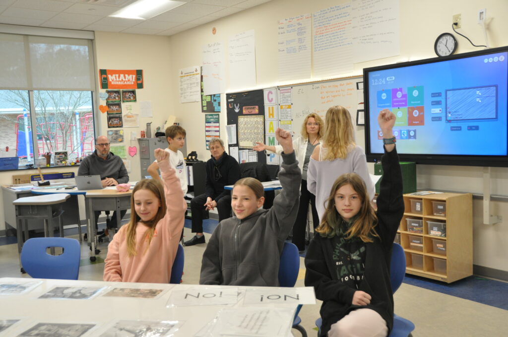 Three 5th grade girls seated at a table in the middle of the room with their arms raised. Serena and David in the background directing the exercise