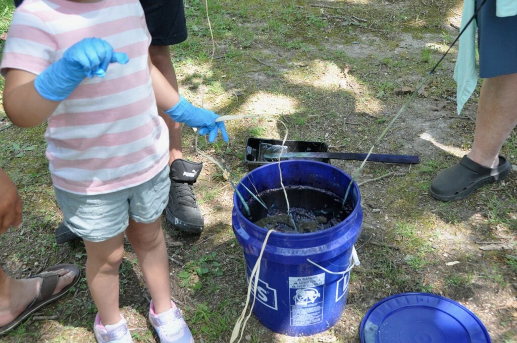 Small child in pink and white striped shirt wiht jean shorts standing next to a bucket of indigo dye with several strings hanging over the edge. Green grass in background.