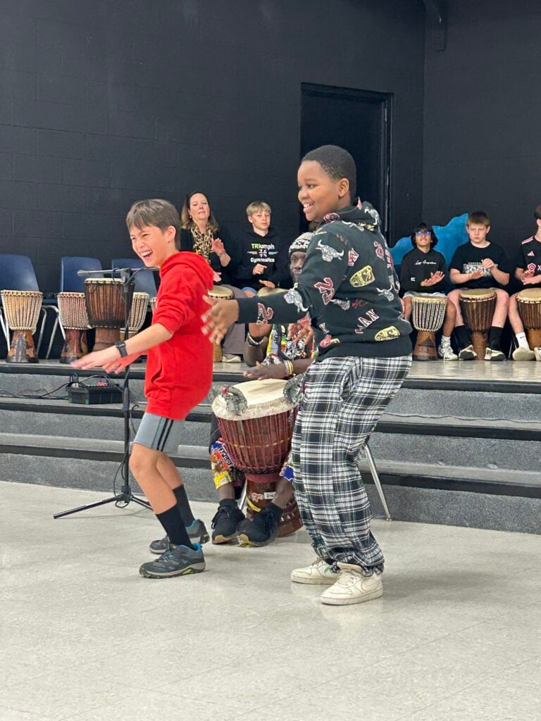 Two boys dancing to djembe drums with student drummers in the background