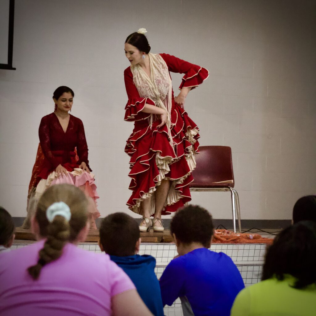 Flamenco dancer in red dress with white trim dancing on stage with children seated on the floor in front