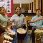 Three girls seated, playing djembe drums while smiling and laughing