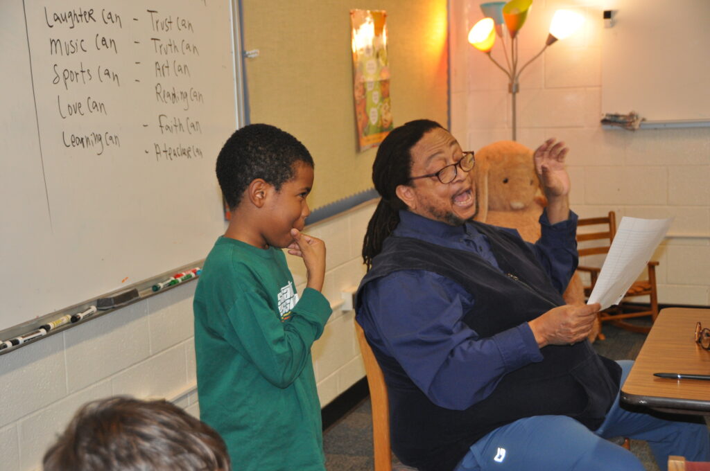 Philip Shabazz mid sentence, seated in a chair reading a poem for a young student standing next to him.