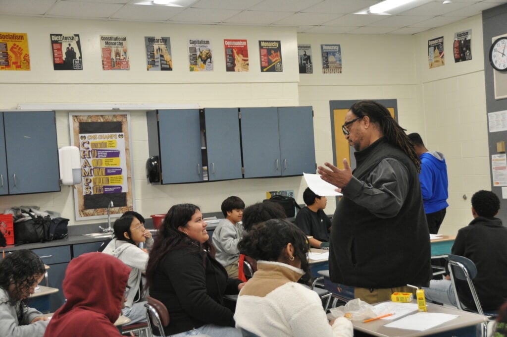 Philip Shabazz stands in front of a classroom, gesturing with his hands while holding a paper, as students sit at their desks.