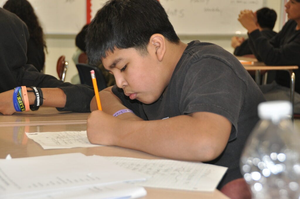A student focuses intently while writing with a pencil on a paper at a desk.