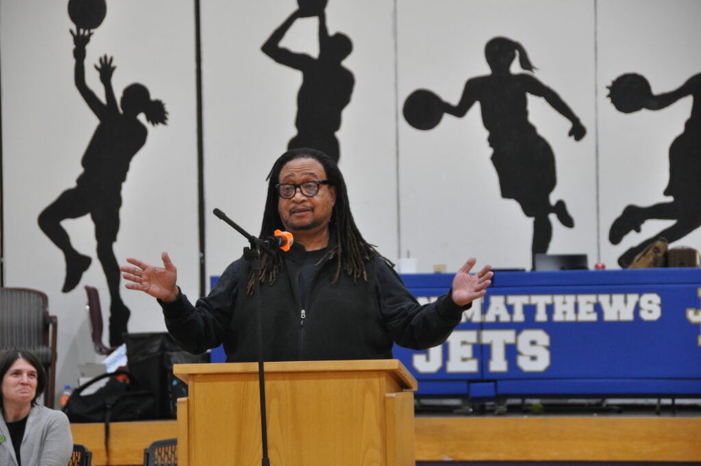 Philip Shabazz gestures with both hands while standing at a wooden podium in front of a backdrop with "MATTHEWS JETS" and silhouettes of basketball players.