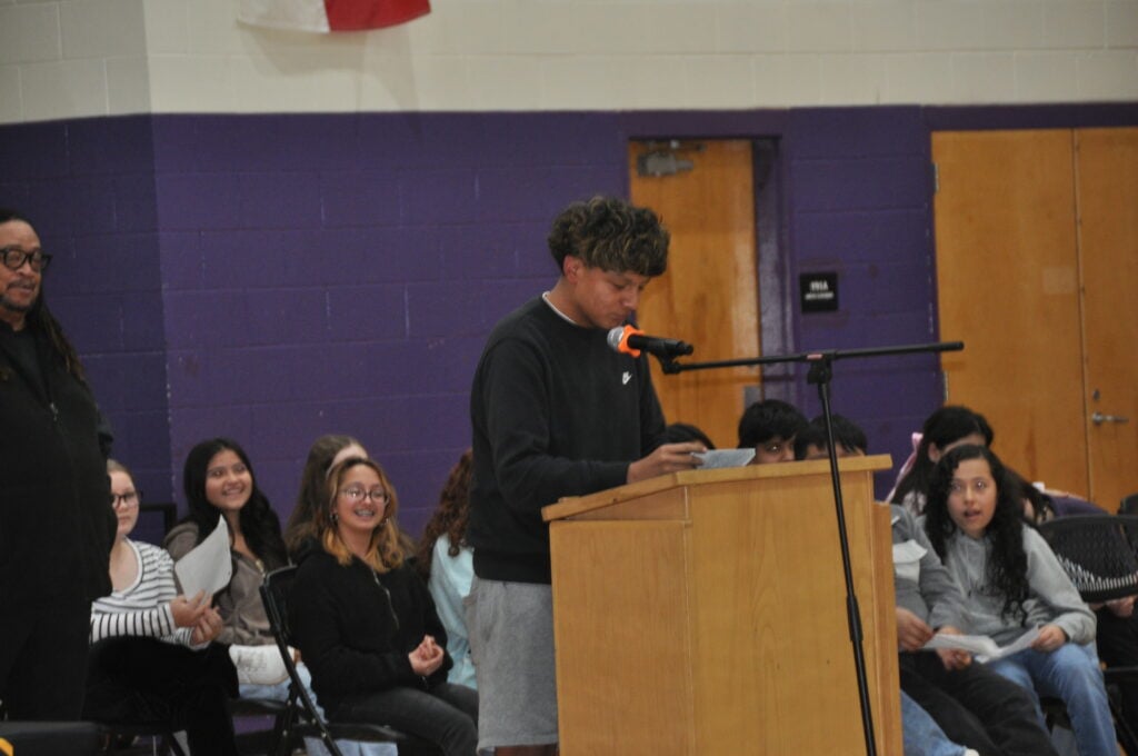 A young person stands at a wooden podium, reading poetry, with an audience of seated individuals behind them. The background features purple walls and a doorway.