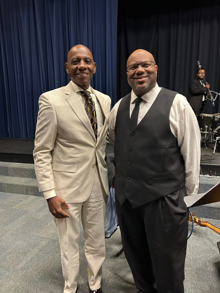 Dr. Anthony Jackson and John Brown in suits stand facing the camera. The man on the left wears a light tan suit with a patterned tie. The man on the right wears a black vest over a white shirt with a black tie. In the background, a band is set up with a double bass player and drums.