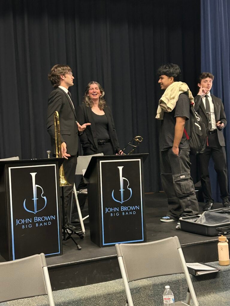 Four people standing on a stage with two music stands labeled "John Brown Big Band". Two are wearing suits, one is wearing casual clothes, and one is wearing a black blazer.
