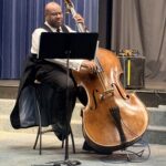 John Brown in a white shirt, black vest, and tie playing a double bass. He is on a stage with blue curtains behind him and a music stand in front of him.