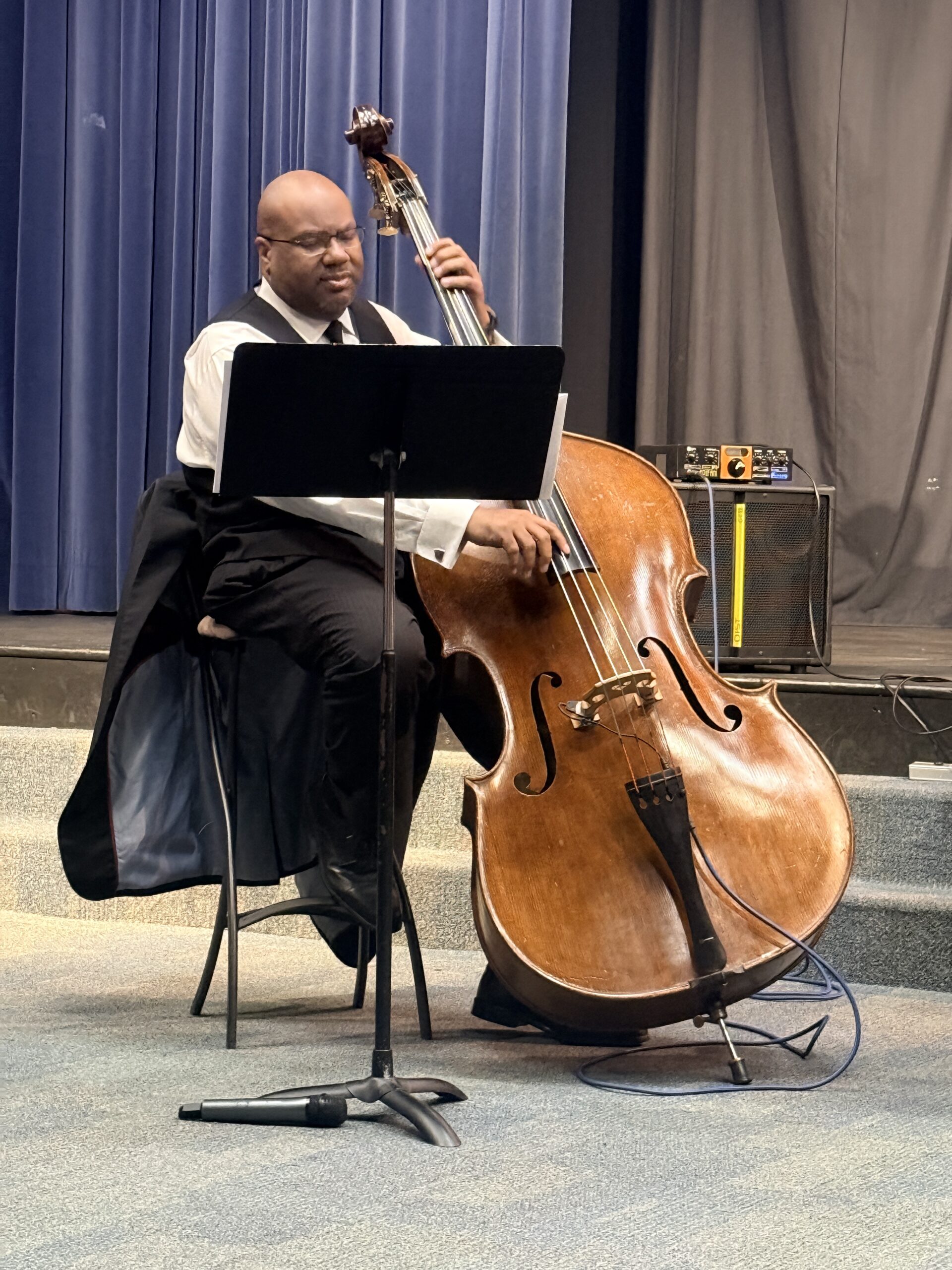 John Brown in a white shirt, black vest, and tie playing a double bass. He is on a stage with blue curtains behind him and a music stand in front of him.