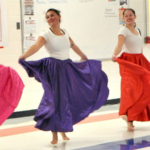 Four dancers in white tops and long flowing skirts—pink, purple, red, and blue—perform in a brightly lit school gym, spinning with arms out while others stand along a wall.