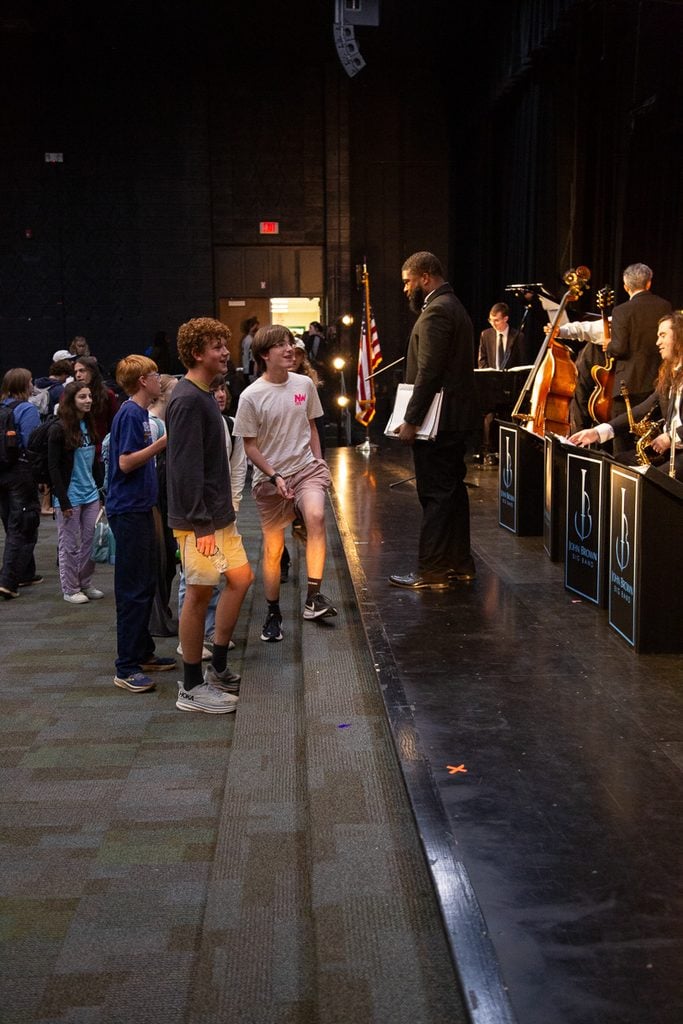 Saxaphone player wearing ablack suit standing on stage, talking with two high school students in t-shirts and shorts with smiling faces.